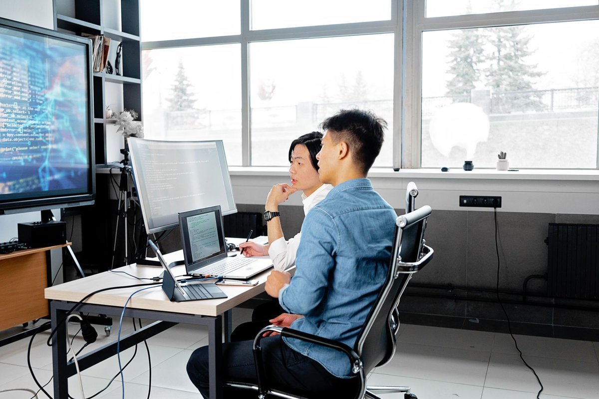 Two colleagues at a shared desk working across monitors showing code
