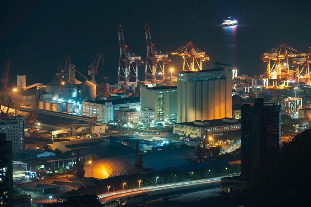 Night industrial port with cranes, silos, and traffic light trails.
