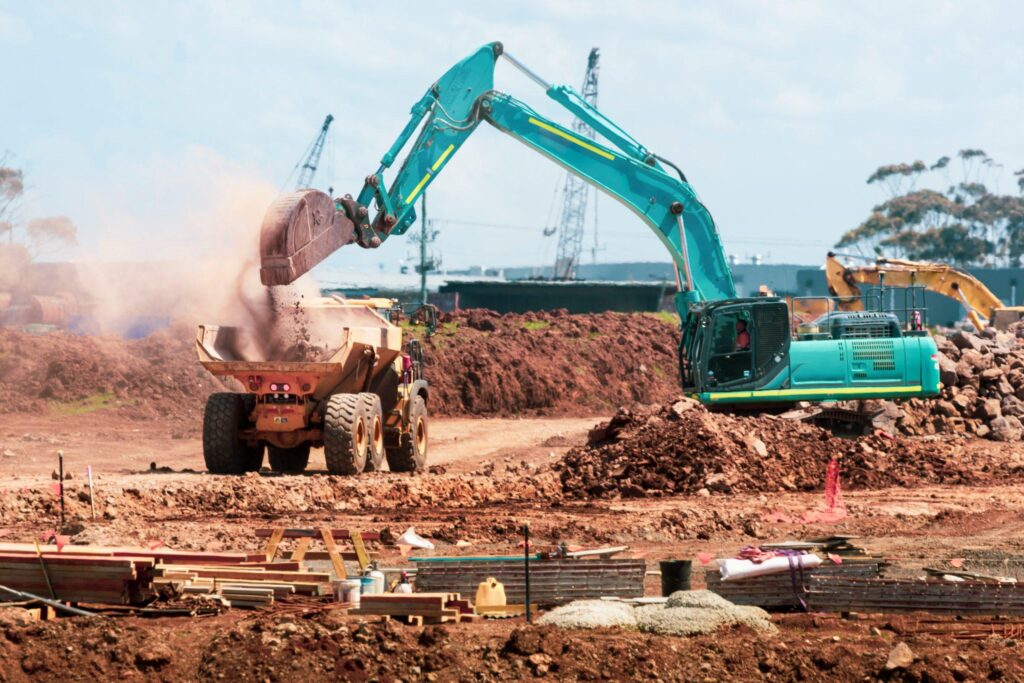 Turquoise excavator loads soil into a yellow dump truck.