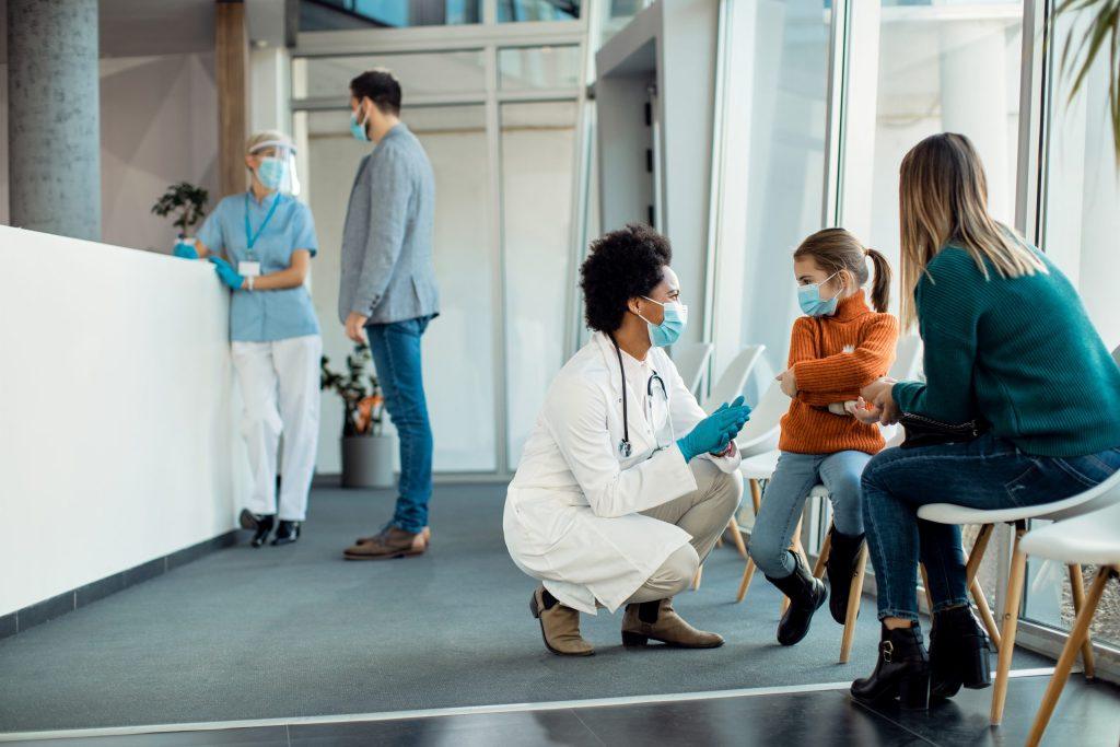 Masked doctor kneels to speak with child and parent in clinic.