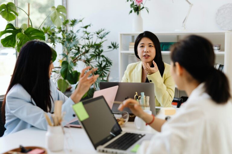 Three women collaborating at a bright office desk with laptops.