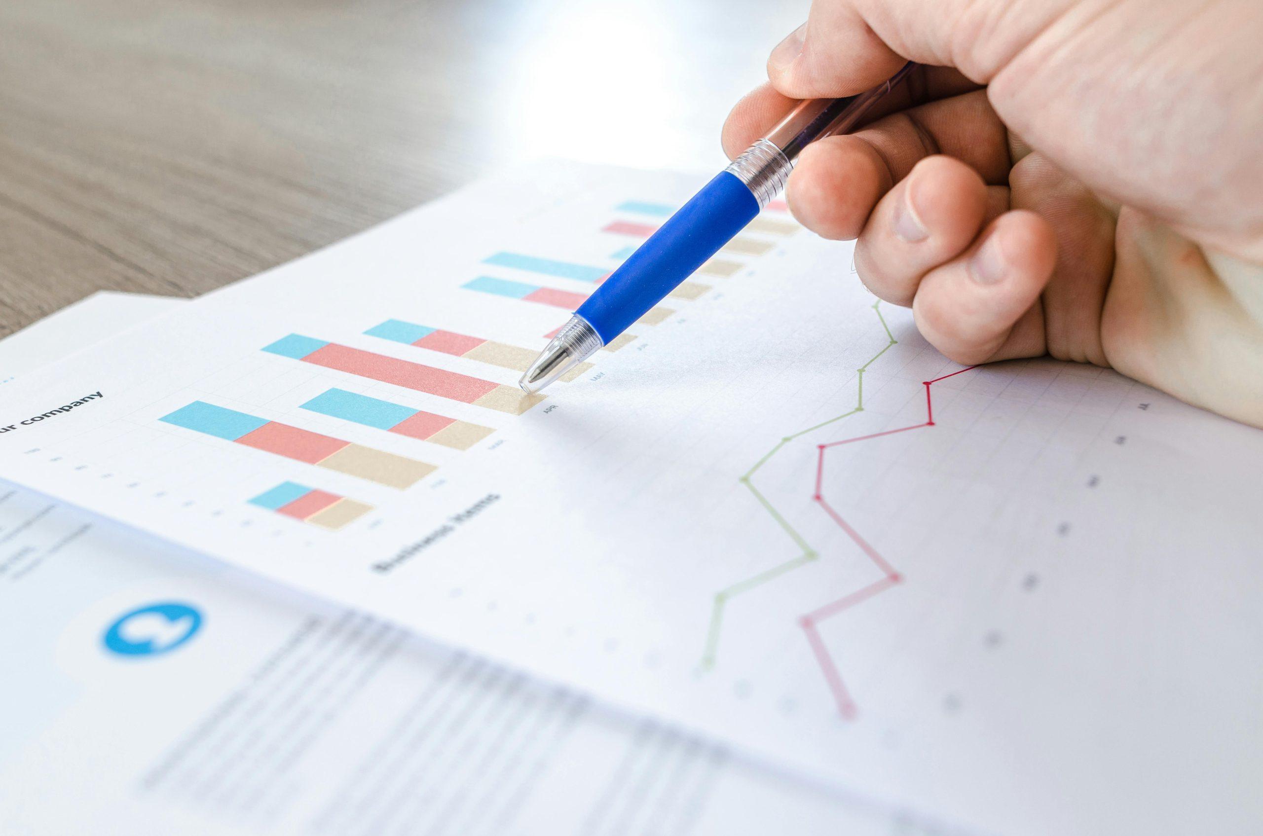 Hand with pen analyzing business charts and graphs.