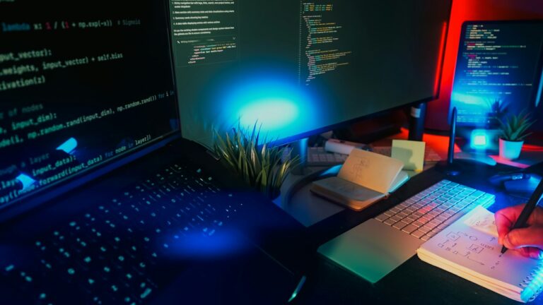 Desk with monitors showing Python code in blue and red light, and a hand sketching a diagram in a notebook