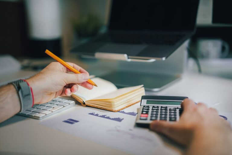 Hands working with a calculator, notebook and printed charts at a desk, with a laptop behind